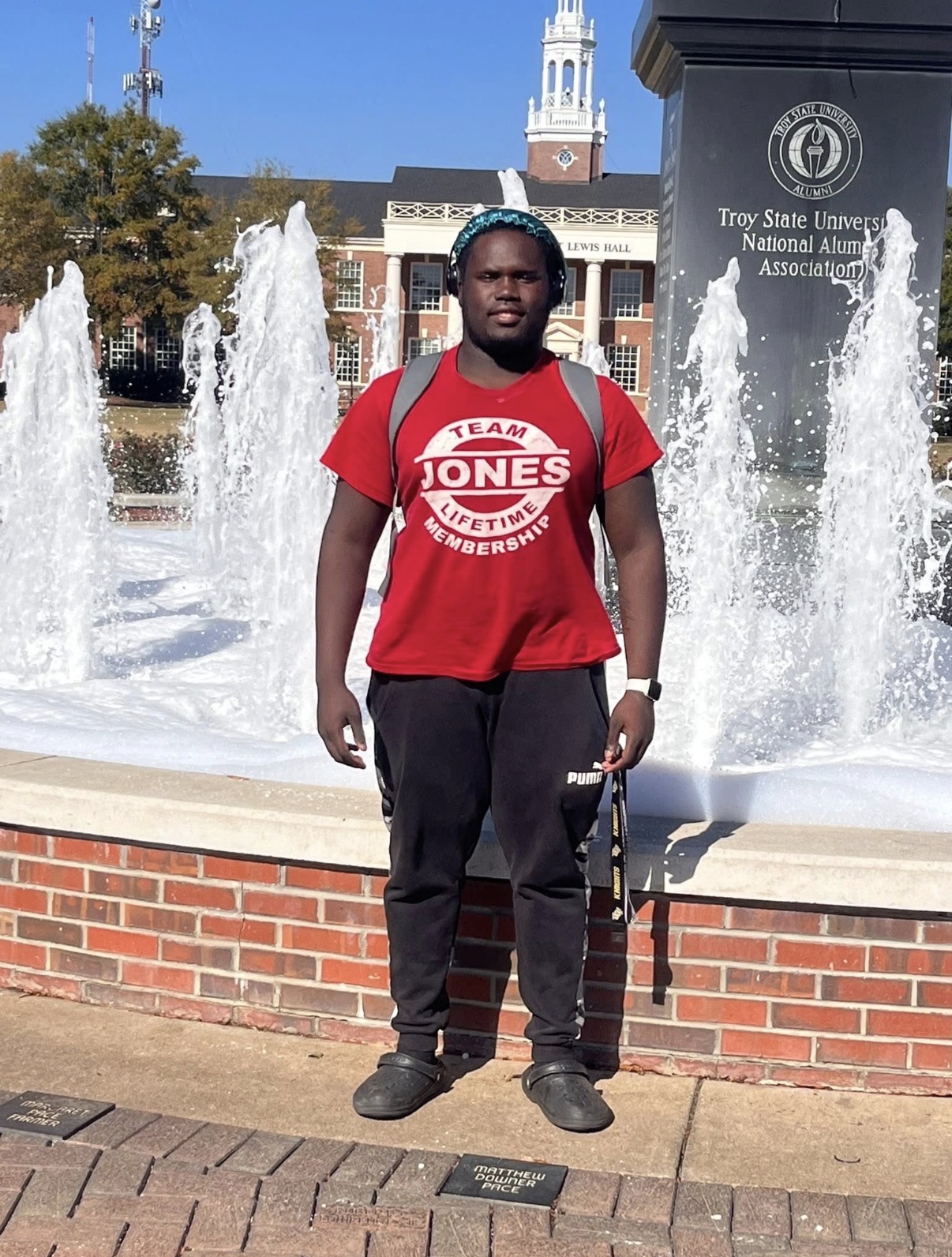 Mahri in a red T-shirt standing in front of a fountain and campus building at Troy University.