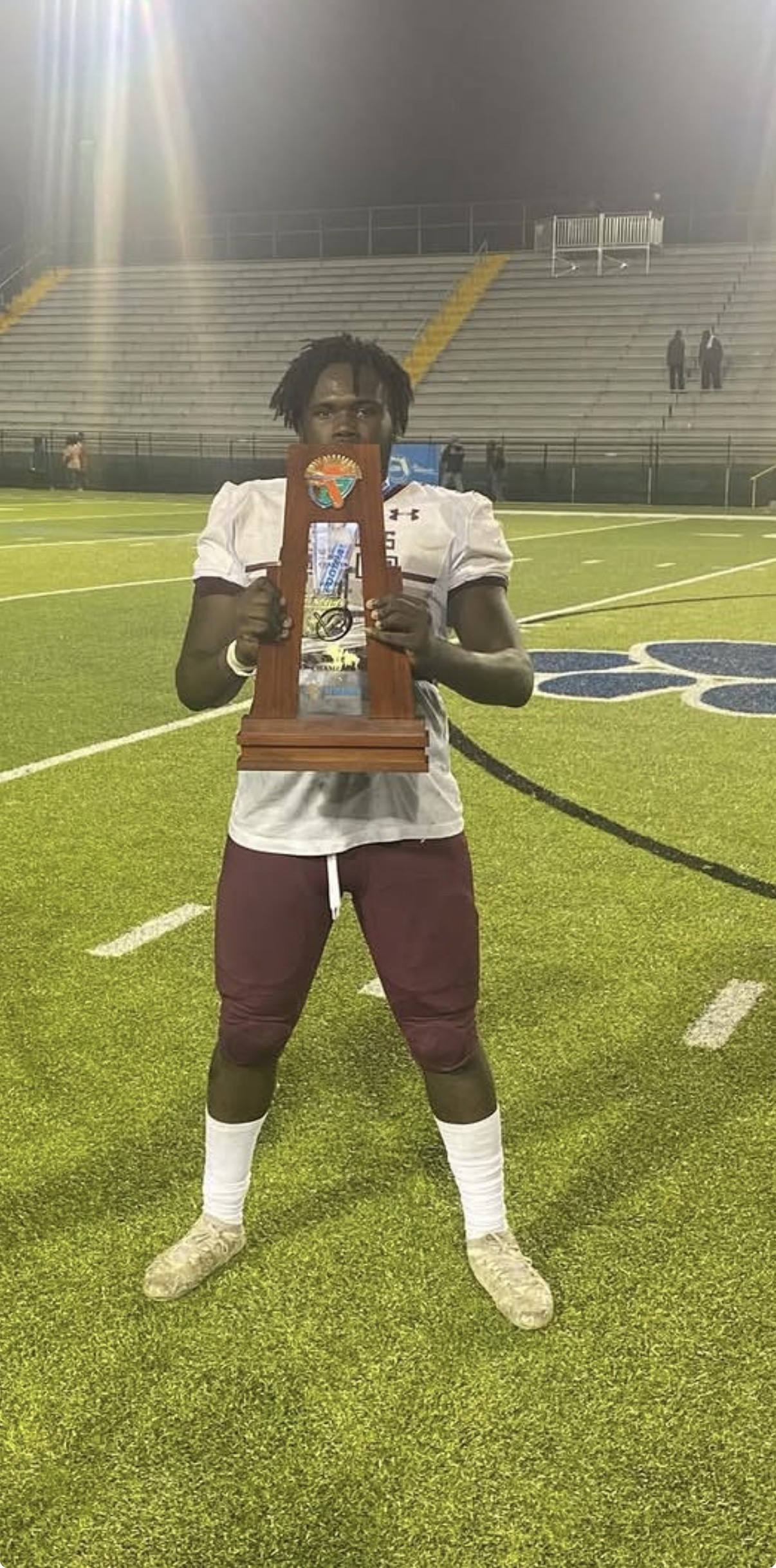 Mahri in a football uniform holding a state championship trophy on a stadium field at night.
